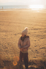 A child girl wearing the hat with a pompon standing on the beach at sunset - 794135560