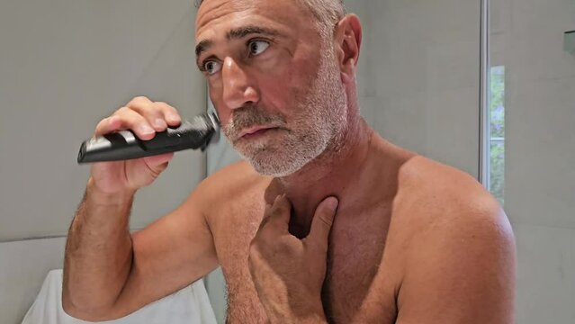 Handsome young unshaven man with towel on shoulders shaving his neck area with modern electric razor trimer