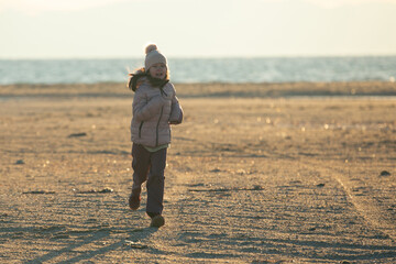 A child girl wearing the hat with a pompon strolling on the beach at sunset