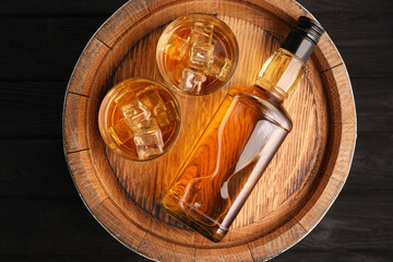 Whiskey with ice cubes in glasses, bottle and barrel on black wooden table, top view