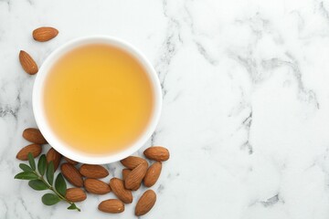 Almond oil in bowl, leaves and nuts on white marble table, flat lay. Space for text