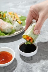 Woman dipping delicious spring roll into soy sauce at grey textured table, closeup