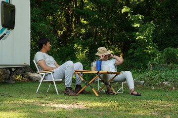 Two Asian male friends with good relations came to relax, have a picnic, sit in the forest and have an RV parked behind them during their camping trip.
