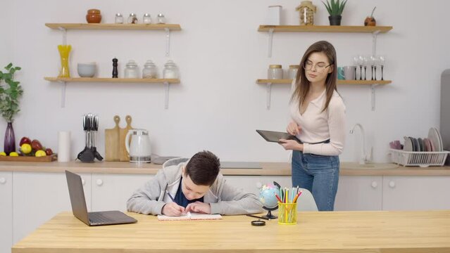Mom helps to son with homework, female standing in kitchen schoolkid takes notes in copybook and laptop accomplish task receives from tutor praises