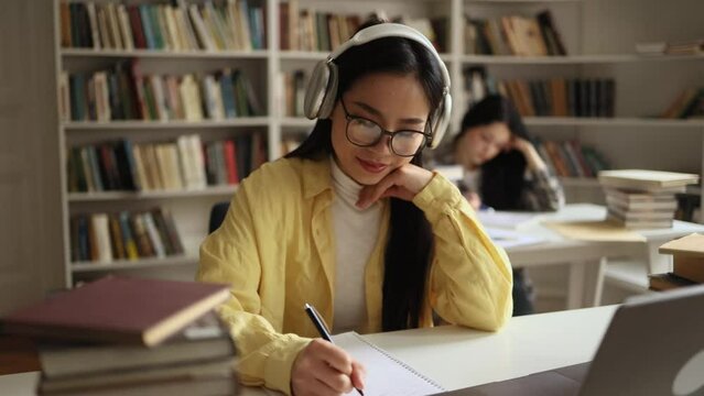 Pretty young asian woman student with headphones listening to music while writes notes for essay study for class preparing for exams test make research for project using books at university library