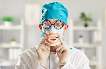 Man dentist, wearing funny glasses and a medical mask, holds a model of a human jaw in a dental...
