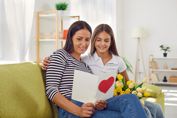 Young mother and daughter sitting on the sofa at home and reading a greeting card together. Happy...