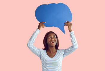 Happy woman holding thinking bubble. Portrait of beautiful smiling young African American girl in casual top standing on pink studio background and holding up empty blue mockup speech cloud