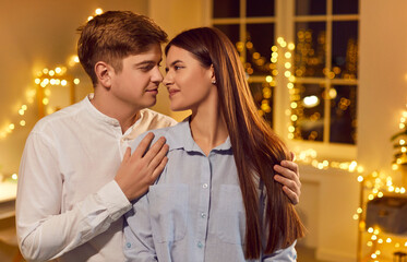 Portrait of cheerful smiling young couple embracing and looking at each other with tenderness standing at home with bokeh background. Happy man hugging his loving wife. Valentines day concept.