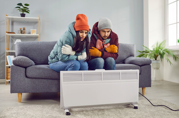 Portrait of a young frozen couple sitting frustrated on the sofa in the living room in winter outerwear and hats at home and trying to keep warm near the electric heater. Heating problems concept.