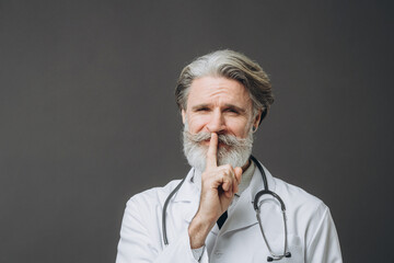 Gray-haired male doctor in white medical coat isolated on gray background raising hand to mouth with silence gesture..
