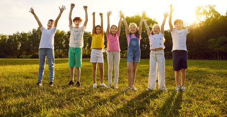 Group of a smiling kids friends raising hands up on green grass in the park standing in a line. Happy children having fun together outdoors on a sunny summer day in casual clothes in the camp. - obrazy, fototapety, plakaty
