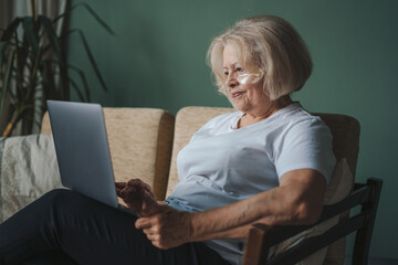 Older woman sitting on sofa with laptop in arms having a conversation with partners. Grandmother using computer alone at home, retired people technology concept