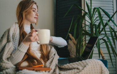 Modern young Caucasian woman having influenza staying at home sitting on sofa writing notes in notebook and drinking tea. Under a blanket, satisfied with the