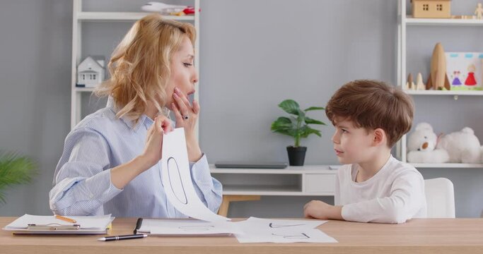 Speech therapist working with child. Language pathologist doing articulation exercises with kid. Female specialist sitting at table with little boy shows him letter O and asks to pronounce sound