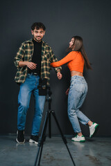 Millennial guy and woman recording content for followers, using cell phone set on tripod, dancing and smiling on black background.