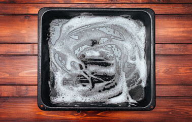 White foam and soap on a black baking sheet that stands on a wooden table. Abstract pattern. The concept of washing the greasy surface of a frying pan.