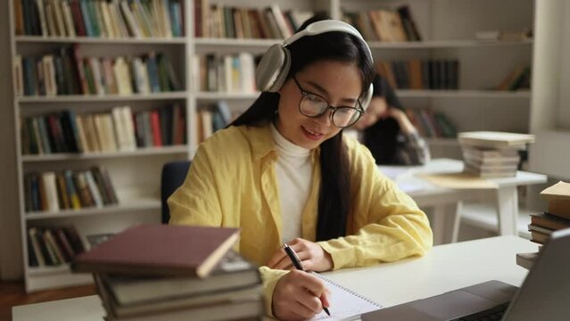 Pretty young asian woman student with headphones listening to music while writes notes for essay study for class preparing for exams test make research for project using books at university library