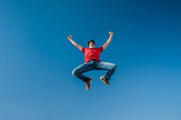 Joyful man in red shirt celebrating with a high jump against clear blue sky