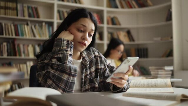 Boring young woman student scrolling feed of social networks products in internet store on smartphone at library workplace Tired exhausted girl look at mobile screen without enthusiasm at university