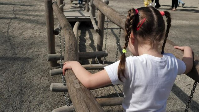 Little preschool girl plays on an interactive playground. Child walks along a rope bridge, overcoming a children's obstacle course. Active recreation, sports and physical development of children.