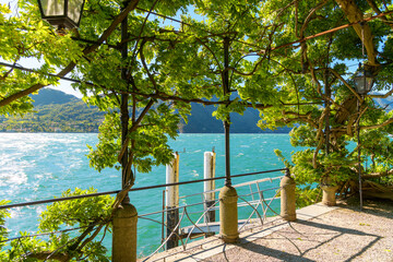 View from a lush plant covered lakefront promenade terrace overlooking the lake at the Italian resort town of Tremezzina, Italy, on the shores of Lake Como.