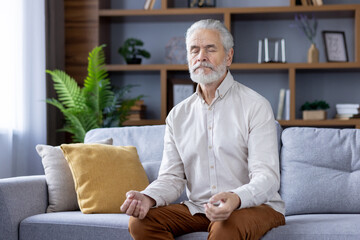 Senior relaxed gray-haired man sitting on sofa at home with eyes closed in lotus position,...