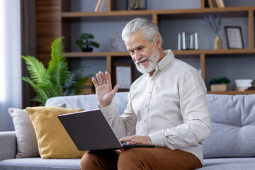 Smiling gray-haired senior man sitting at home on sofa with laptop and talking on video call to...