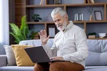 Foto op Plexiglas Smiling gray-haired senior man sitting at home on sofa with laptop and talking on video call to camera, waving and greeting © Tetiana