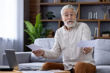 Foto op Plexiglas Portrait of a worried senior gray-haired man sitting on a sofa at home and working on a laptop, checking bills and received receipts, looking upset at the camera and throwing up his hands © Tetiana