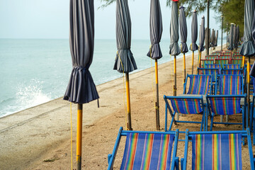 Waves from the sea are splashing stone stairs wall next to umbrella beach and many blue chair on sandy beach under pine tree in the morning at a seashore, South Cha-am Beach, Phetchaburi, Thailand.