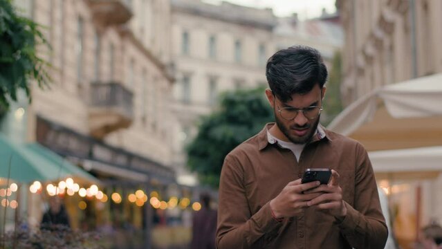 Unhappy Indian Arabian ethnic male traveler man guy tourist exploring city route travelling outdoors. Businessman stroll abroad street looking mobile phone gadget cellphone walk town using online map