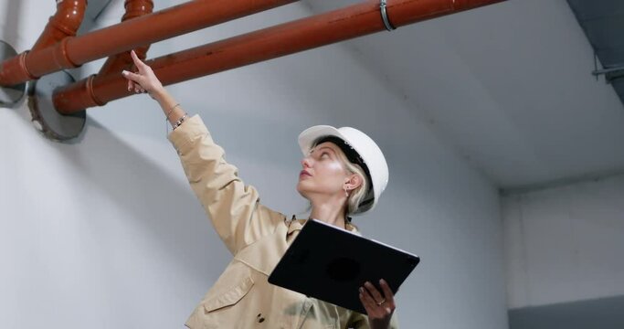 Female inspector in helmet examines fire hydrant system comparing with plan on tablet. Lady waves hand indicating middle-level condition