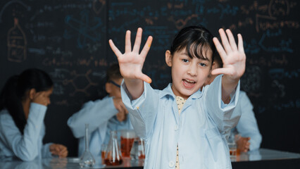 Cute asian scientist waving hand to camera while diverse group doing experiment. Young schoolgirl...