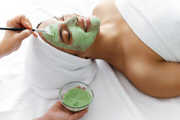 Close-up of process of therapist applying green face mask on half face of a beautiful brazilian or hispanic young woman. Skincare concept. Procedure of applying a clay mask to the female face