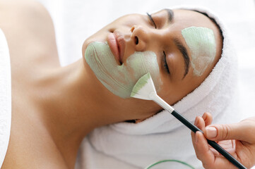 Close-up of beauty procedure, therapist applying green face mask on half face of a beautiful brazilian or hispanic young woman. Skin care concept. Facial treatments, facial cleansing