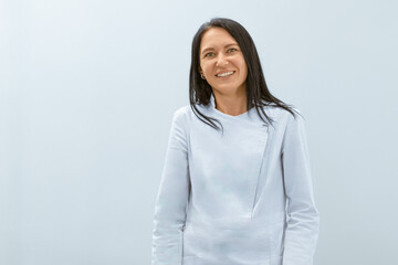 A female massage therapist, smiling warmly, stands confidently in professional attire, her black hair cascading down her shoulders, facing the camera with approachability.
