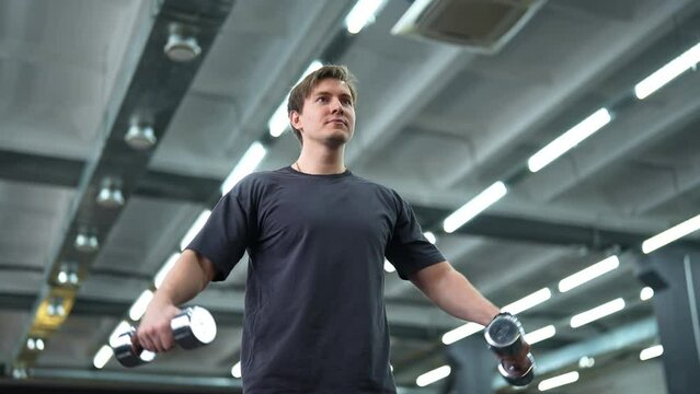 Low-angle portrait of motivated beginner sportsman doing side lateral raises with dumbbells at gym. Active handsome man exercising to stay fit and slim at sport club. Shooting in slow motion.