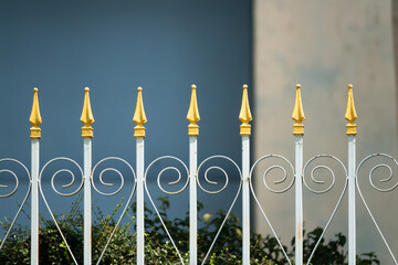 A fence with gold spikes on it. The fence is white and has a blue background