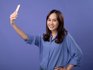 Joyful Asian woman taking a selfie with her smartphone, dressed in a casual blue blouse, against a vibrant purple background