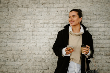 Portrait of a chic woman with cellphone smiling and standing near wall