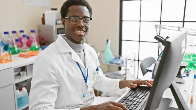 African american male scientist working at a computer in a lab, surrounded by scientific equipment.
