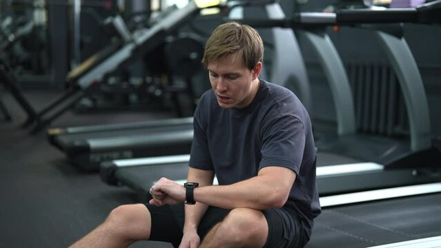 Tired beginner sportsman taking break at sport gym sit on treadmill. Portrait of exhausted sweaty runner hard breathing after cardio exercise running workout. Weary man sit on fitness treadmill relax