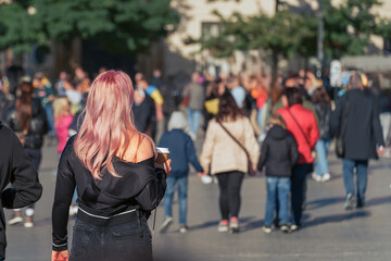 Pink-haired millennial girl with a glass of coffee or tea in her hand on a city street stands out among the noisy city crowd. Modern youth urban lifestyle and individuality