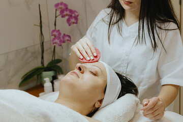 A girl cosmetologist cleanses the patient s face with a scrubbing sponge.