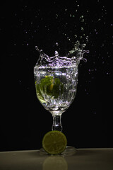 a slice of lemon falling into a glass cup with clear water causing splashes. Isolated on dark background.