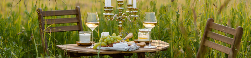 Beautiful romantic outdoor wedding decor in a field. Table decorated with burning candles, flowers. Wineglasses with white wine. Sunset, summer, golden hour. Perfect surprise date for loving couple