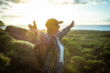 Carefree senior caucasian woman with hat and backpack in outdoor excursion standing with open arms...