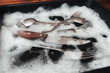 A knife, fork and spoons in the detergent foam on a black oven-tray. Washing dishes concept.