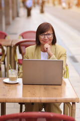 Mature stylish confident businesswoman in eyeglasses and suit works on laptop drinks coffee at European cafe outdoors. Concept of remote work from public place, digital freelance and modern lifestyle